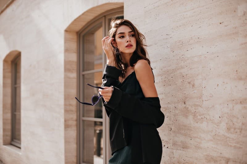 cute young girl with dark wavy hairstyle bright makeup silk dress black jacket holding sunglasses hands looking away against beige building wall 1.jpg